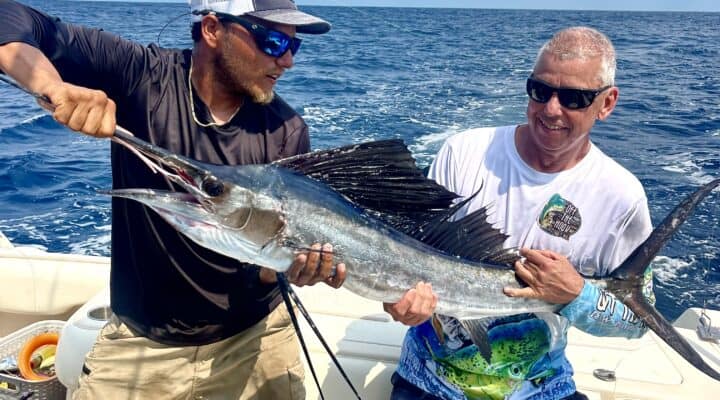 Vibrant image of two men on a boat in Belize holding a large sailfish caught during a fishing charter, showcasing thrilling sportfishing experience in Belize Reef waters.