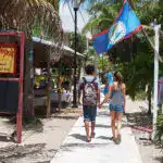 People walking on a sandy pathway with tropical plants, restaurants, and a Belize flag in the background, offering a scenic view of Belize Reef Charters and Belize's coastal attractions.
